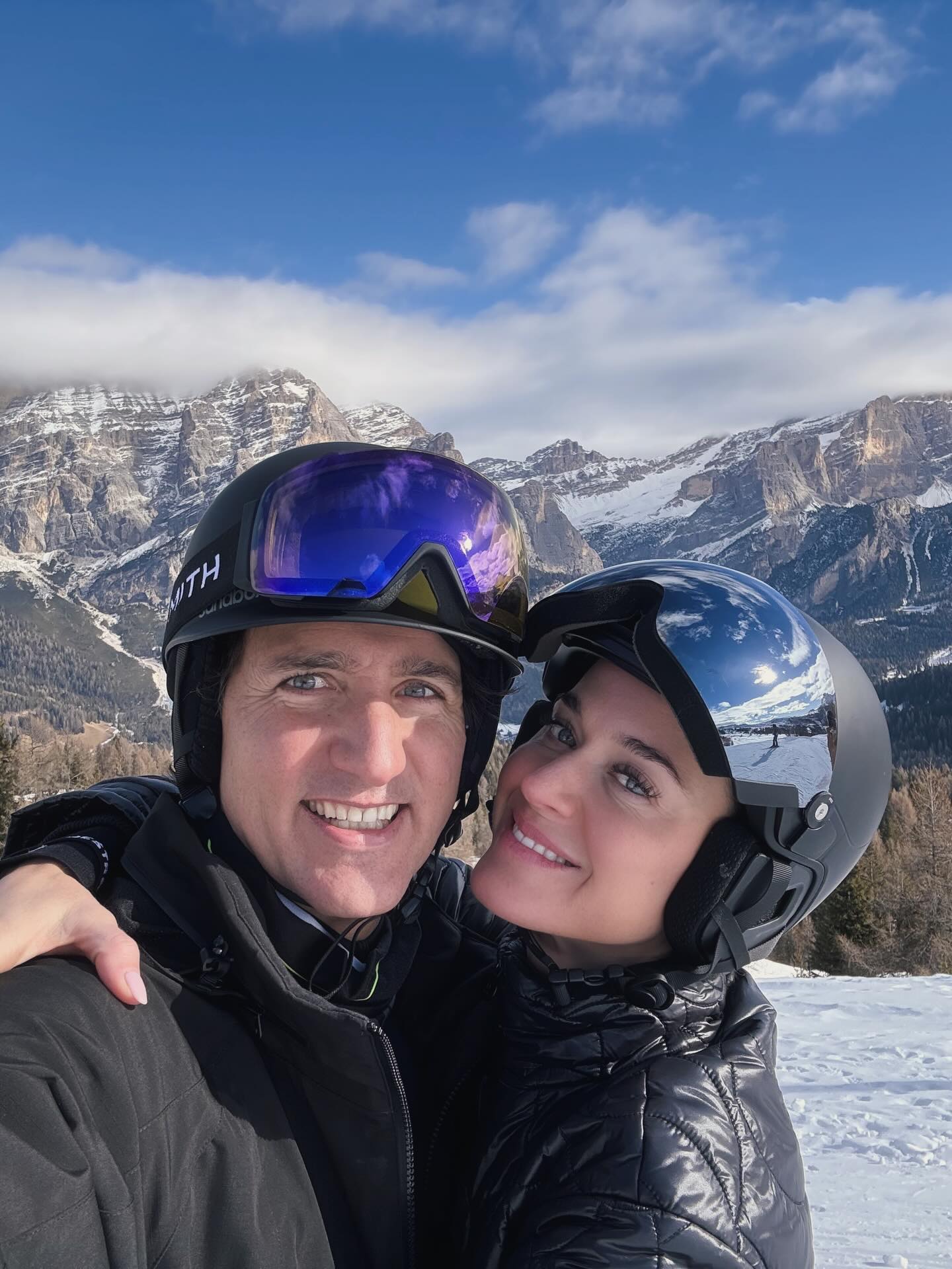 Katy Perry and Justin Trudeau wearing ski helmets and goggles, smiling in front of snowy mountains.