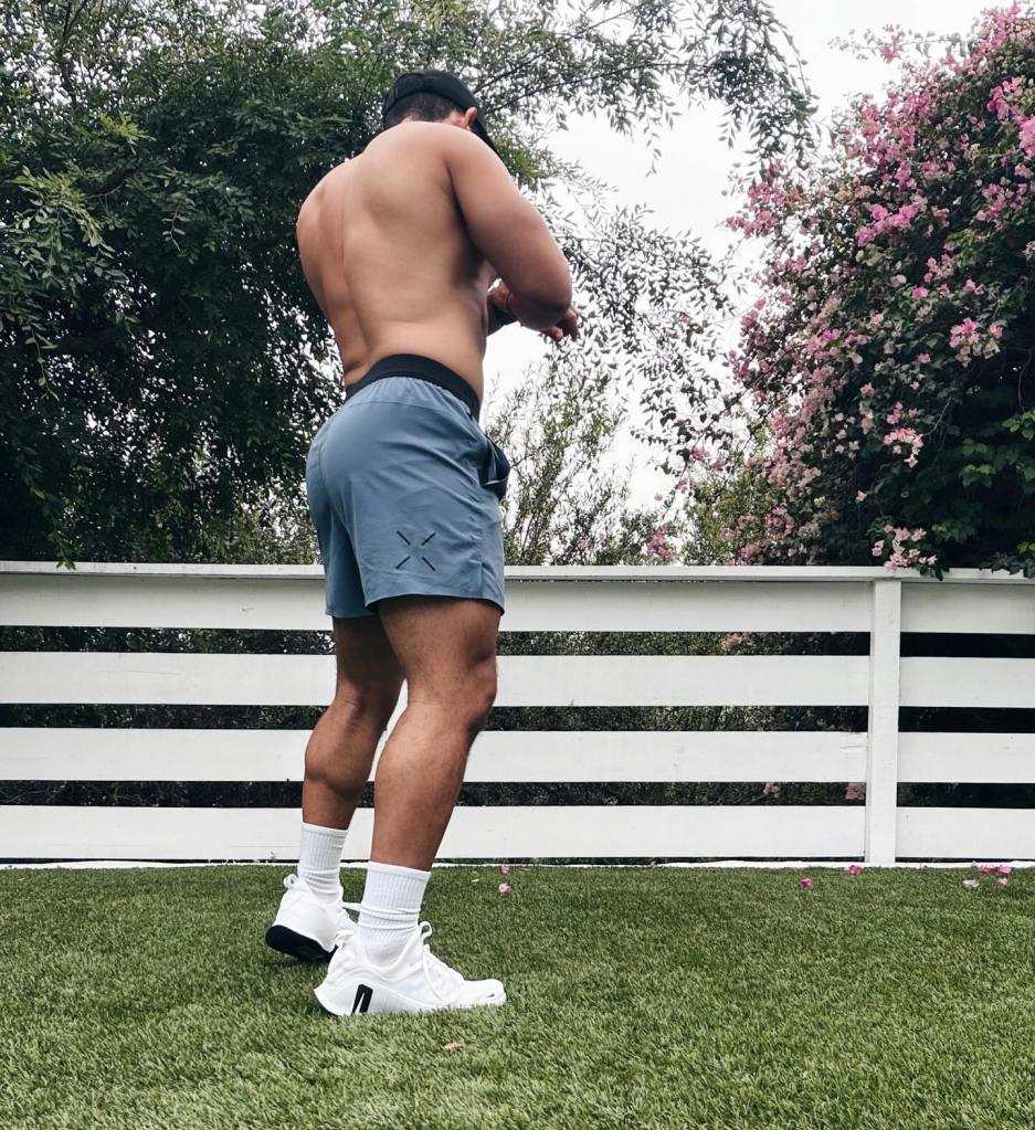 Tahj Mowry facing away from the camera, shirtless and wearing blue shorts, on green grass.