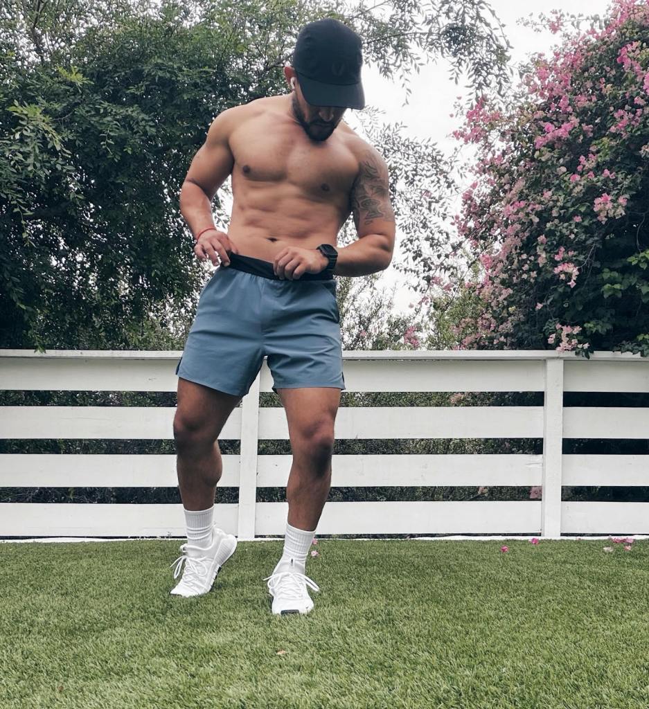 Tahj Mowry shirtless, wearing a cap and shorts, standing on grass.