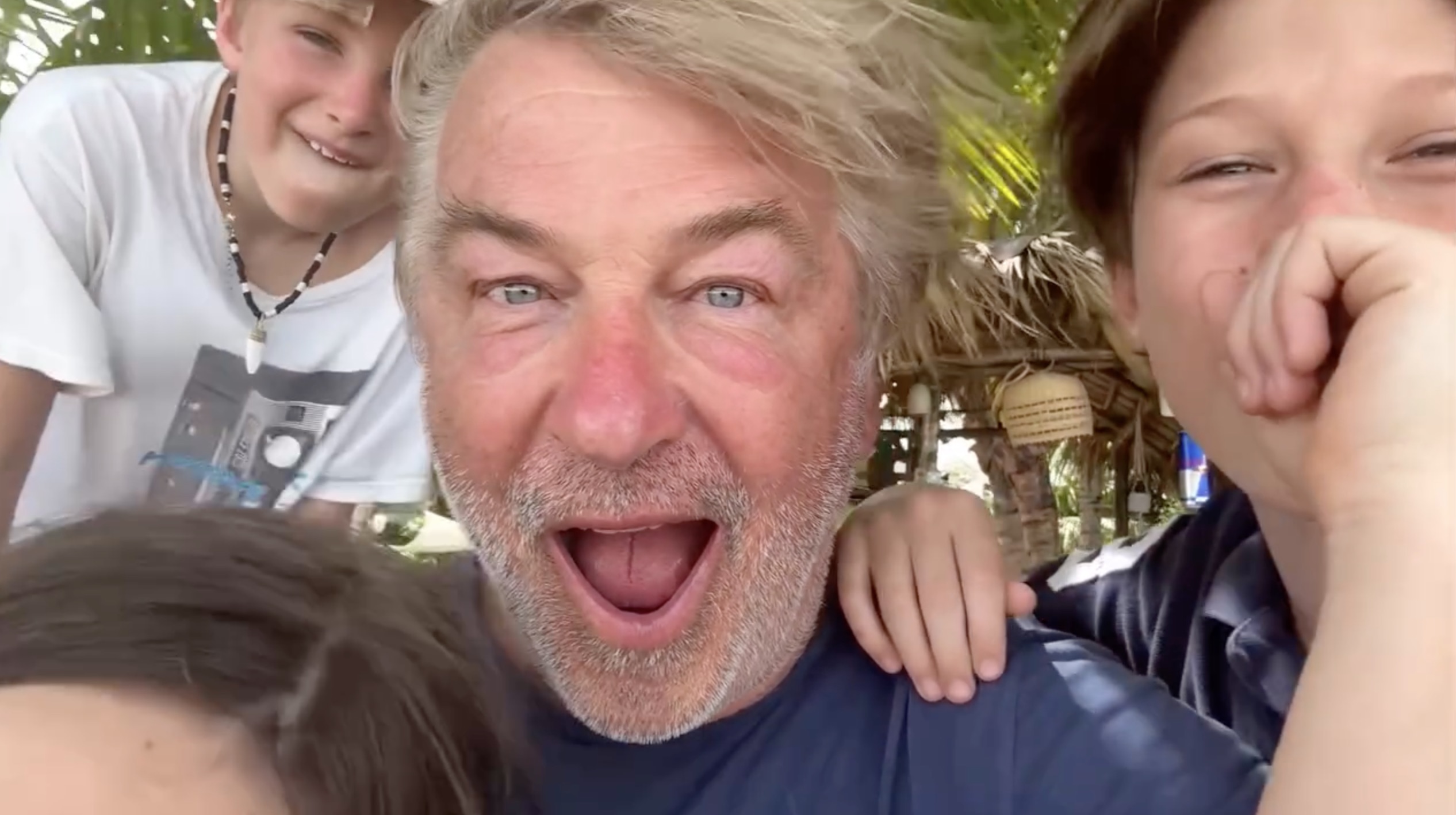 Alec Baldwin with two children, one smiling in the background and another covering their mouth, with Baldwin's mouth open in surprise.