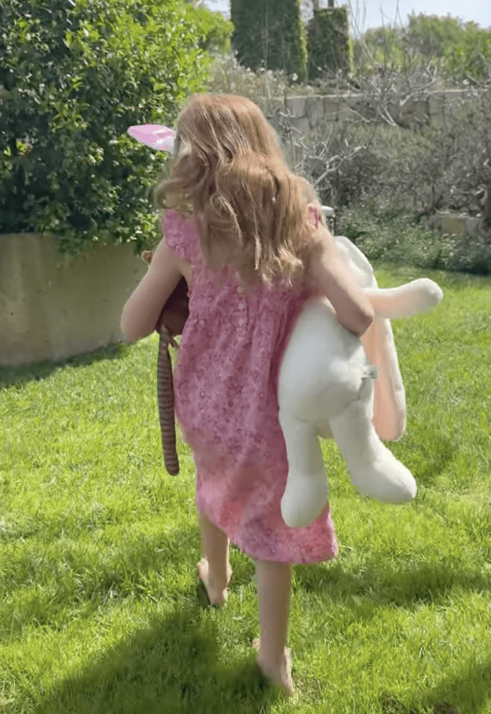 A young girl wearing bunny ears and a pink dress walks barefoot on grass while carrying a plush rabbit and another stuffed animal.