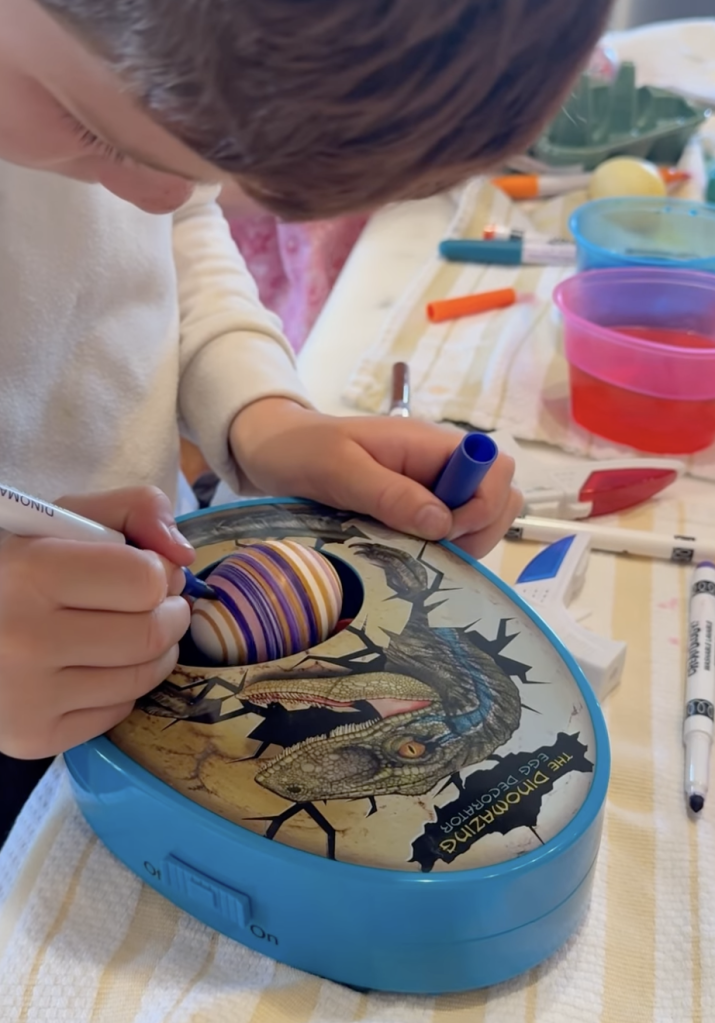 A child decorating a striped Easter egg in a dinosaur-themed egg decorator.