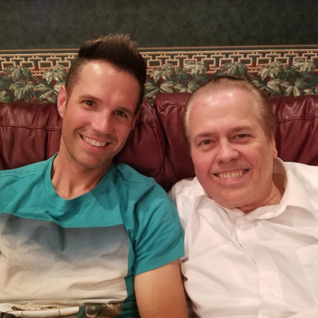 Alan Osmond and his son sit on a couch smiling.