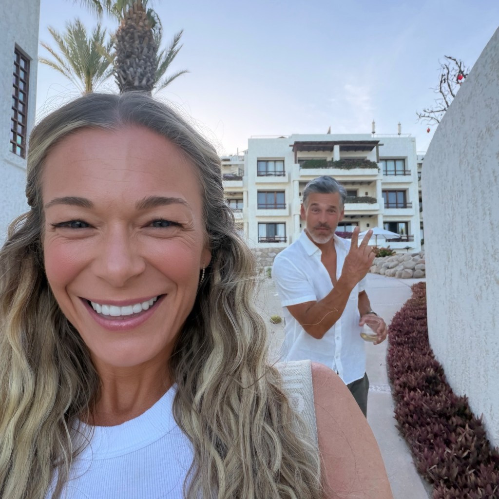 LeAnn Rimes smiling in a selfie with Eddie Cibrian holding a drink and making a peace sign in the background.