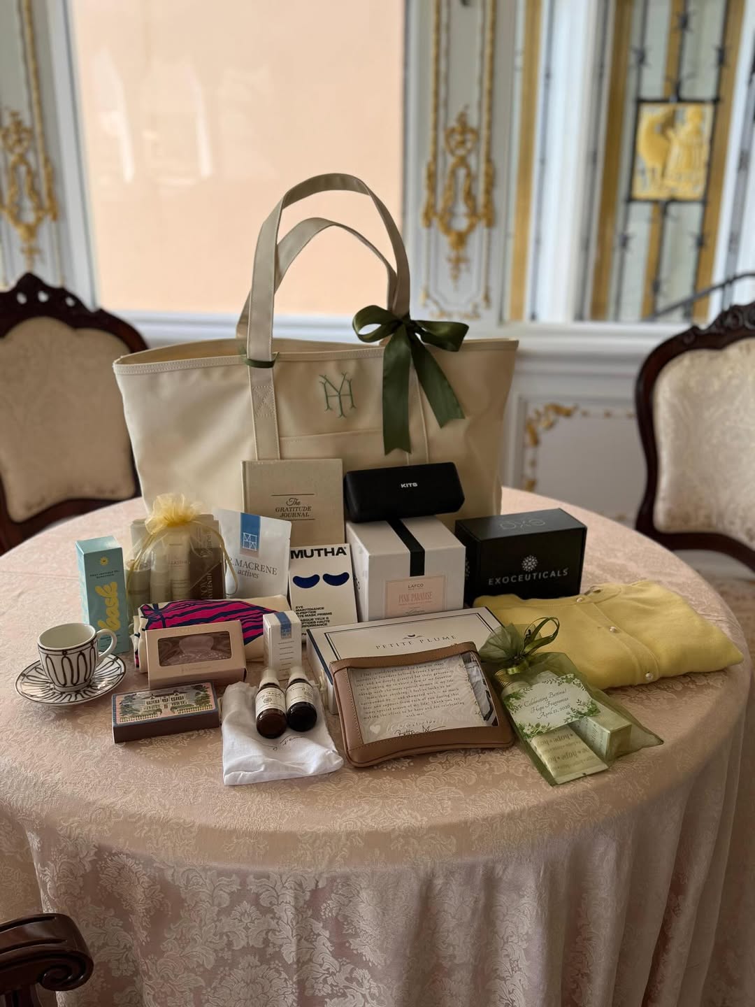 Gift bags and gifts displayed on a round table, including a large cream-colored tote with a monogram, various beauty products, a small yellow sweater, and a patterned teacup.