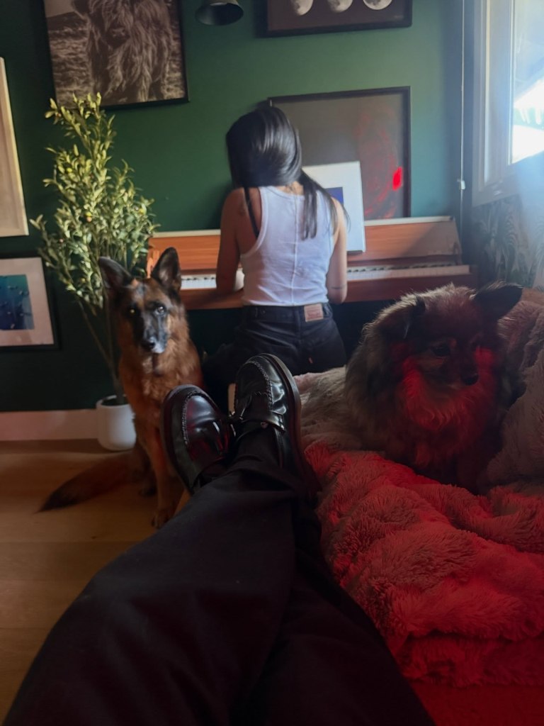 Janel Parrish playing piano with Sasha Farber's feet in foreground, and two dogs looking on.