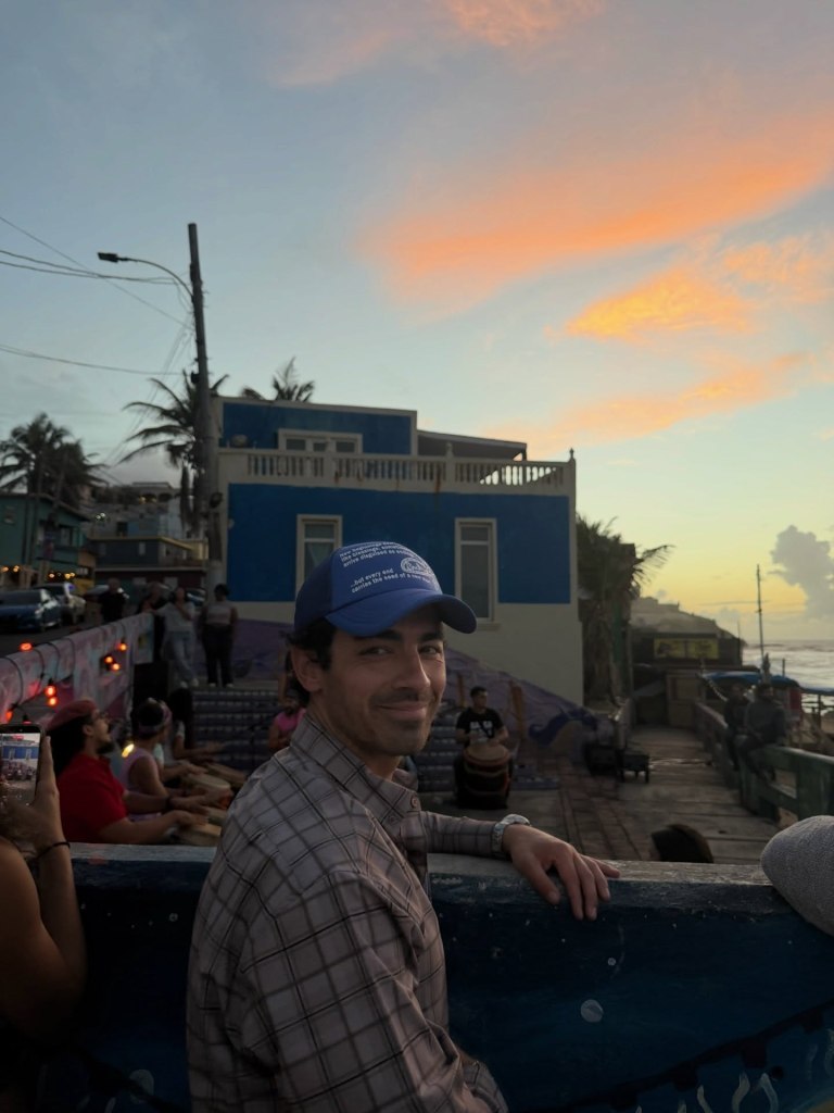Joe Jonas wearing a blue cap and plaid shirt, smiling at the camera with a sunset over the ocean in the background.