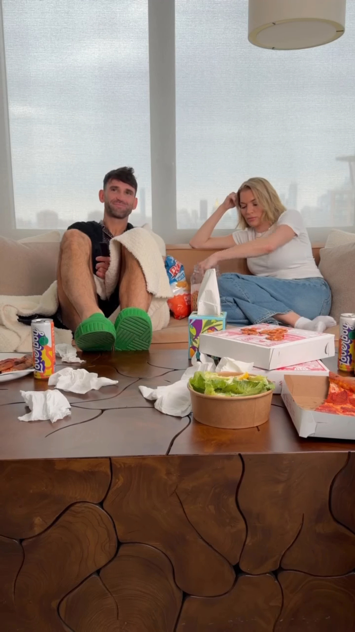 Carl Radke and Lindsay Hubbard lounging on a couch with pizza, chips, and salad on a coffee table.