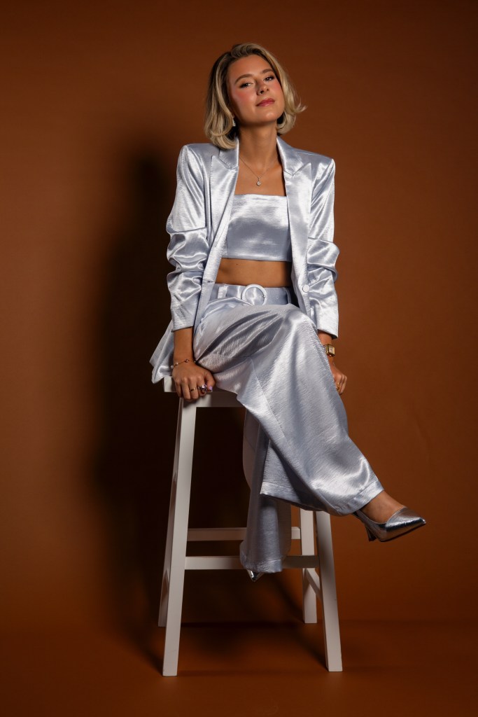 Olivia Levin, author of The Story of Us, in a shiny silver outfit, sitting on a white stool against a brown background.