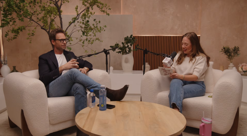 Patrick J. Adams and a woman sitting in armchairs, speaking into microphones on a podcast set.