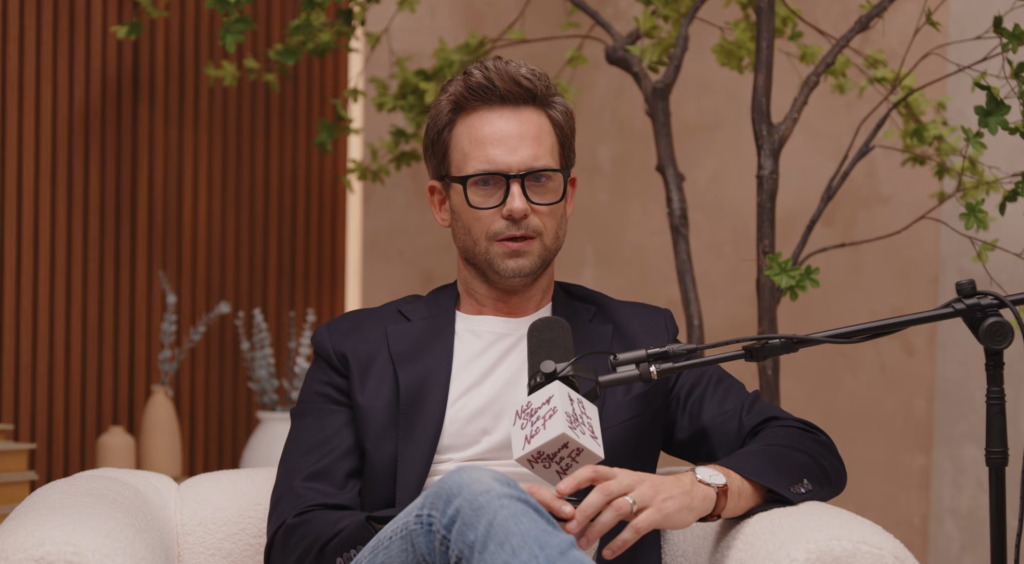 Patrick J. Adams sitting in a chair, wearing glasses and a suit jacket, speaking into a podcast microphone.