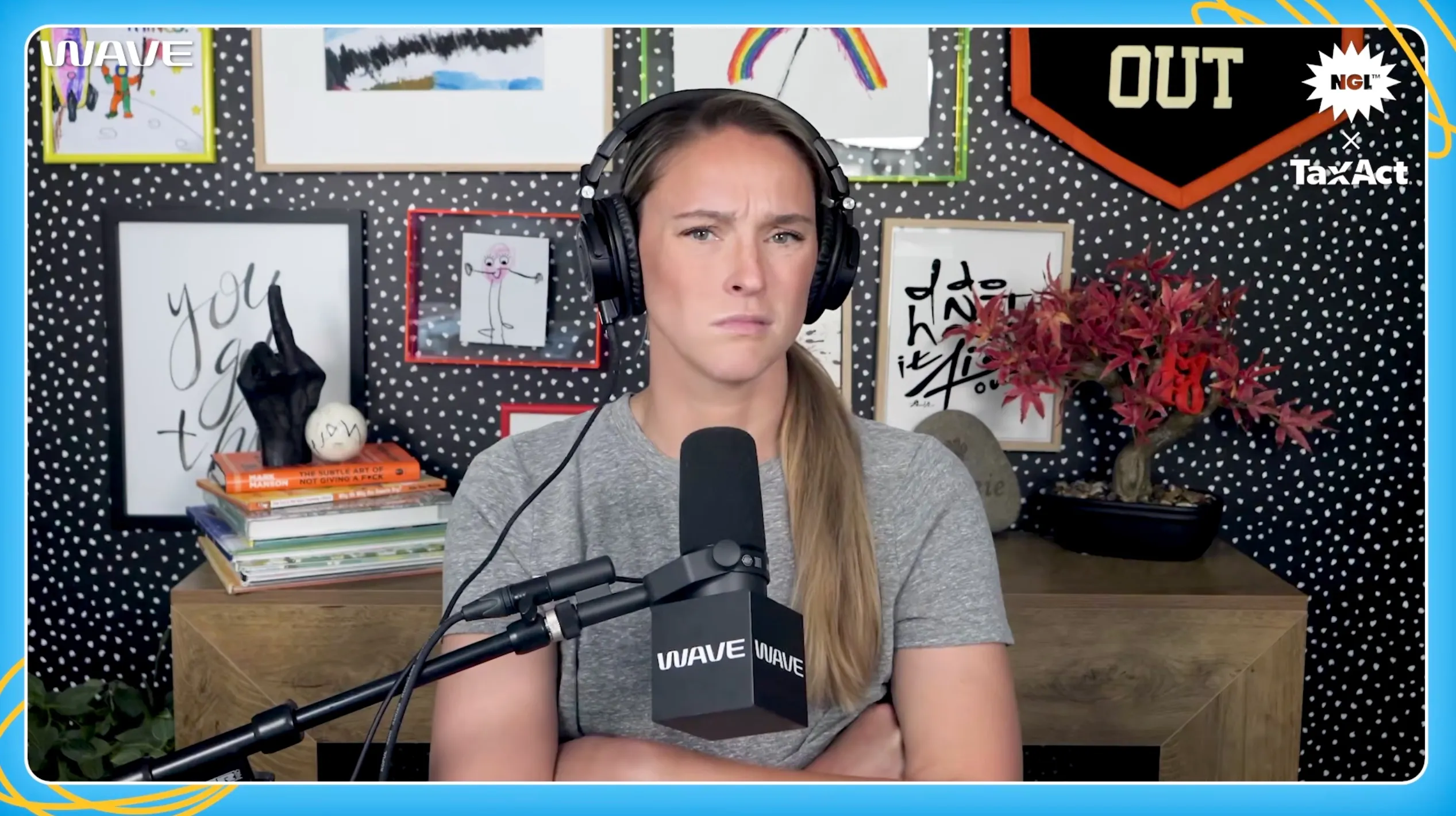 A woman wearing headphones and a gray t-shirt looks seriously at the camera while sitting in front of a microphone.