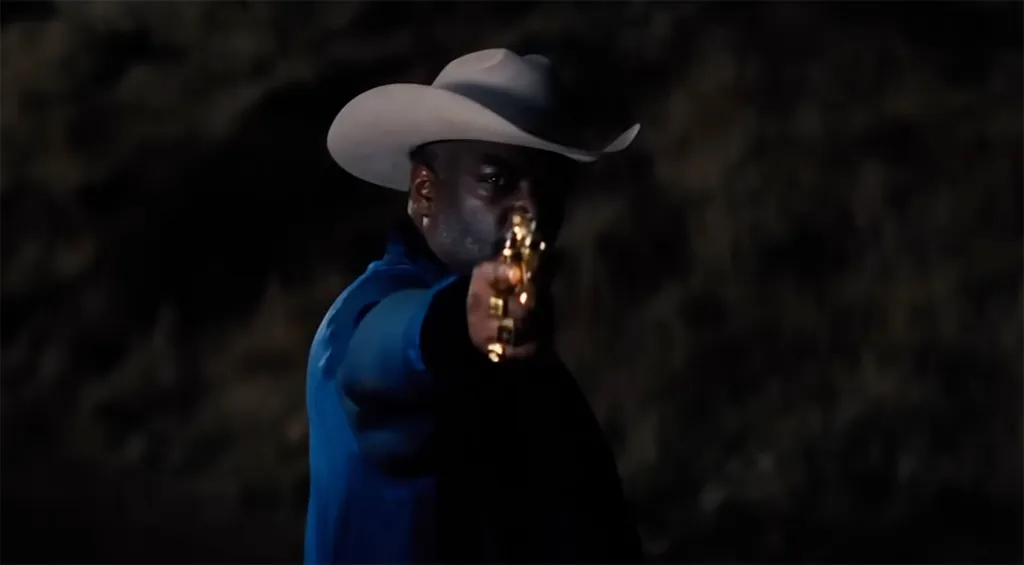 Adewale Akinnuoye-Agbaje in a cowboy hat aiming a golden gun.