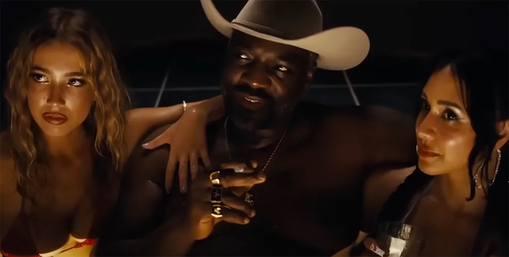 Adewale Akinnuoye-Agbaje in a cowboy hat smoking a cigar, with two women on either side of him.