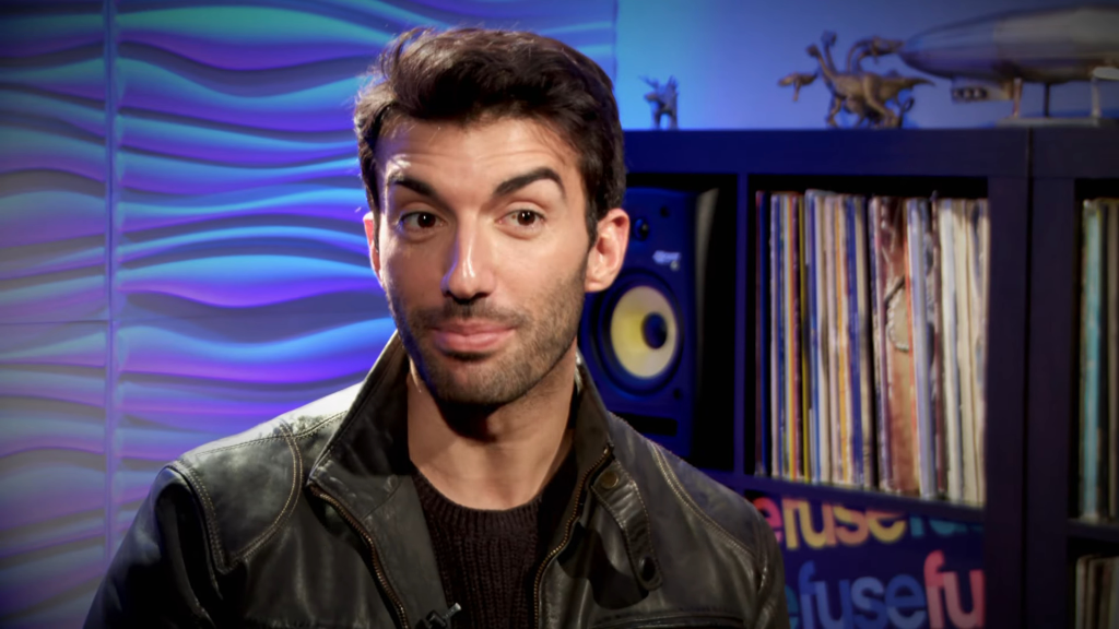 Justin Baldoni looking directly at the viewer with a slight smile, wearing a dark jacket, in front of a blue textured wall and a shelf with records and figurines.