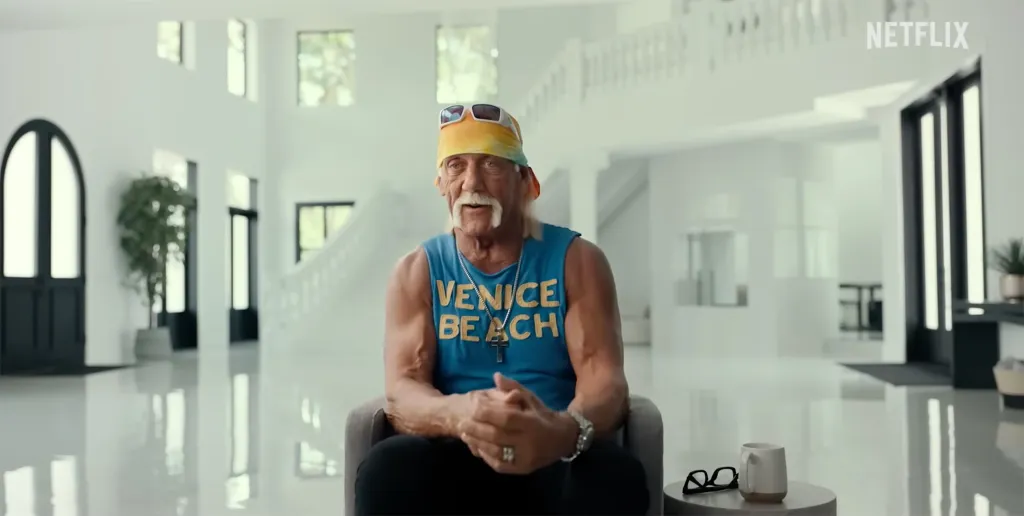 Hulk Hogan in a blue tank top and bandana sitting in a modern white room.