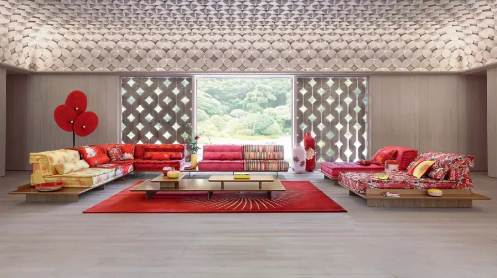 A brightly lit room with a patterned white ceiling, white wooden walls, a light wooden floor, and patterned red couches on either side of the room.