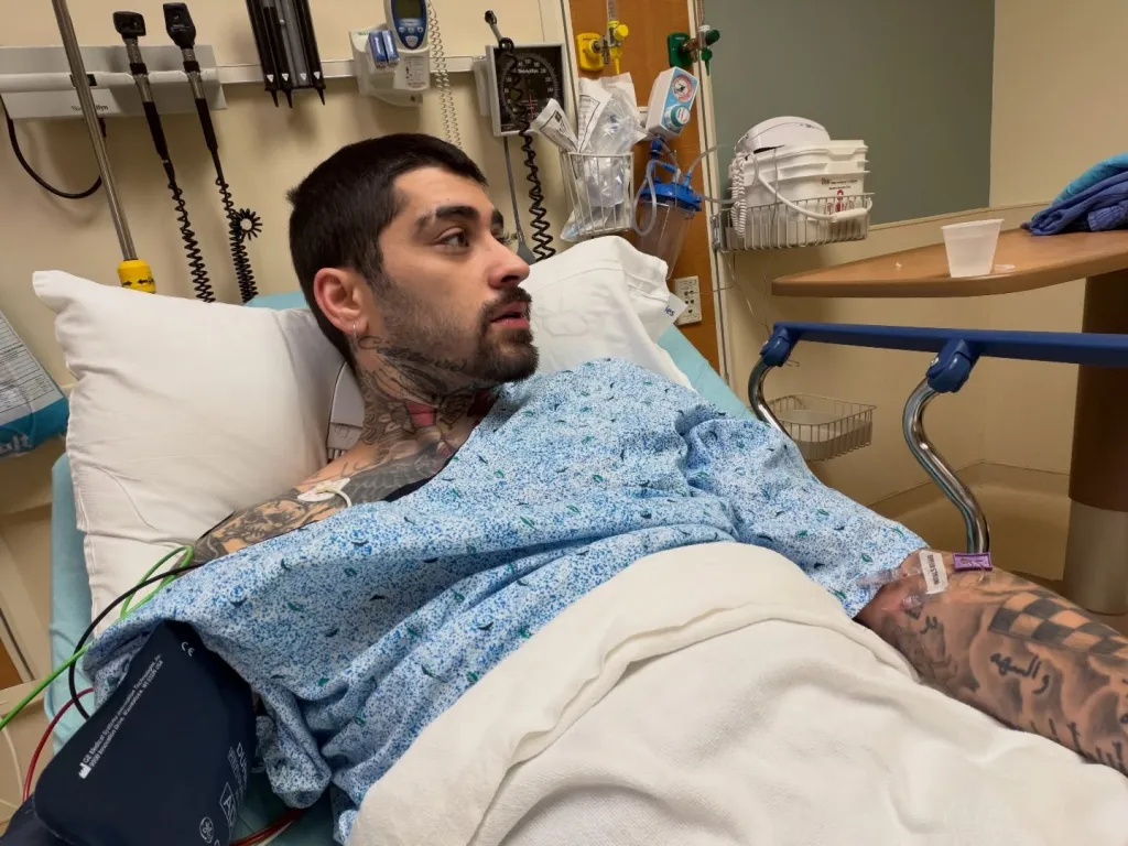 Zayn Malik lying in a hospital bed with an IV drip and a blood pressure cuff on his arm.