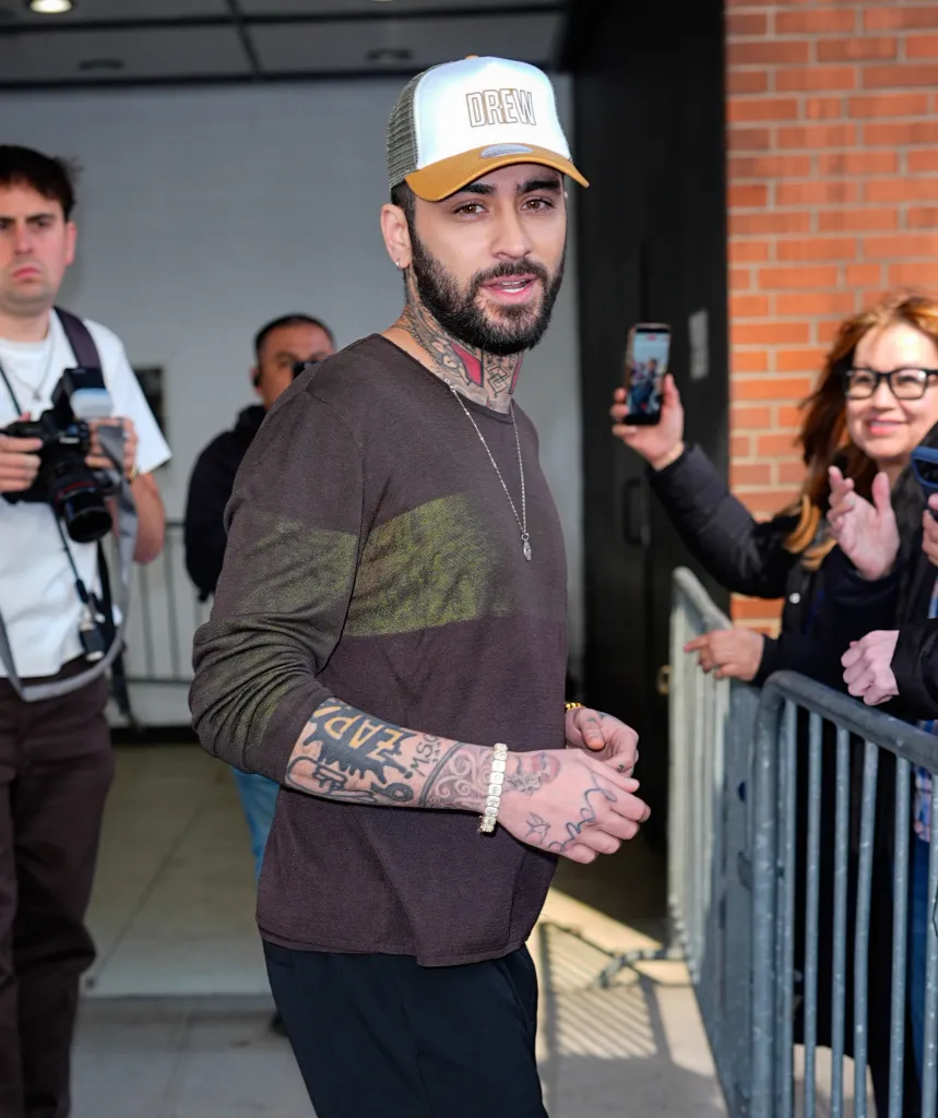 Zayn Malik wearing a white and brown baseball cap, a brown shirt, and a gold bracelet while outside in New York City.