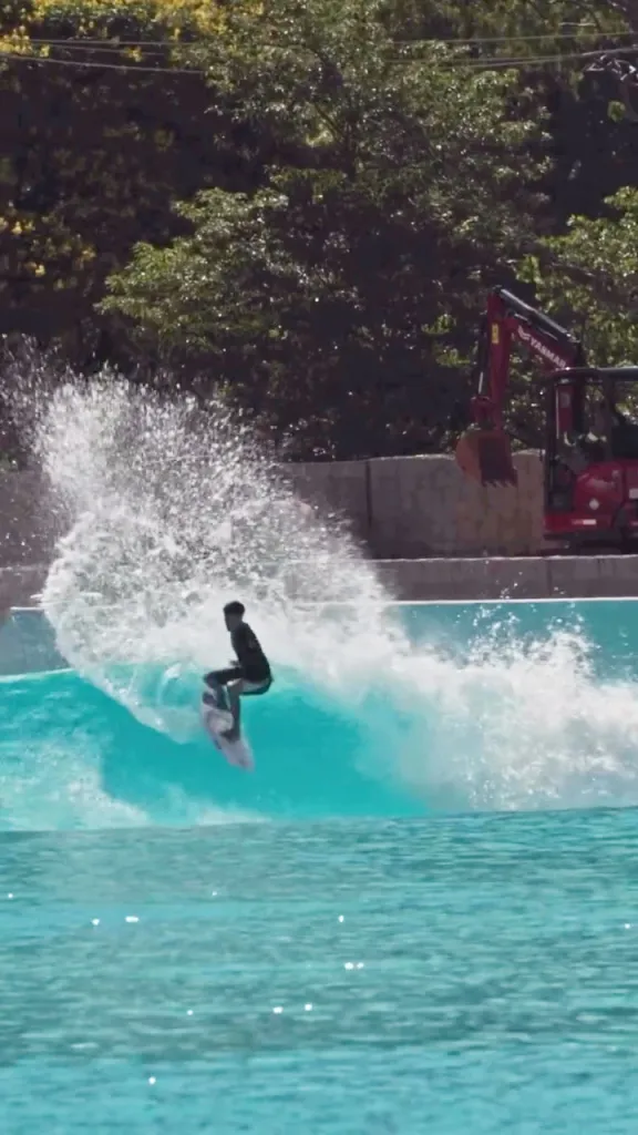 A surfer rides a large wave in a wave pool, with a red excavator and lush green trees in the background.