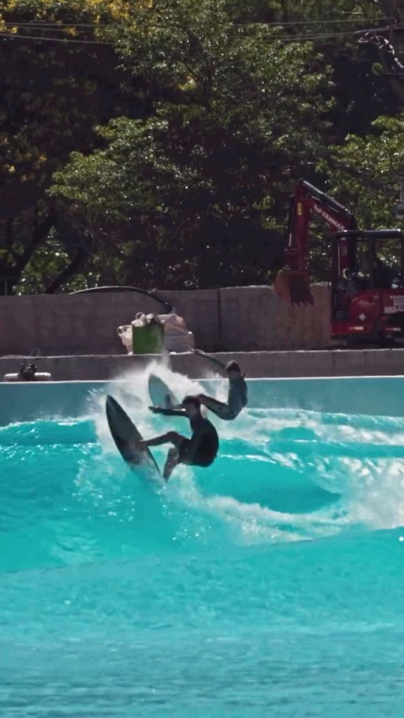 Surfer riding a wave in an artificial wave pool with another surfer falling in the background.