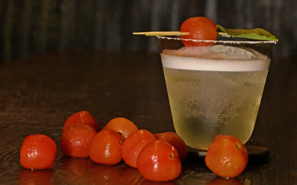 A drink in a glass with a foamy top and a red cherry tomato on a skewer, with more cherry tomatoes on the dark wooden surface.