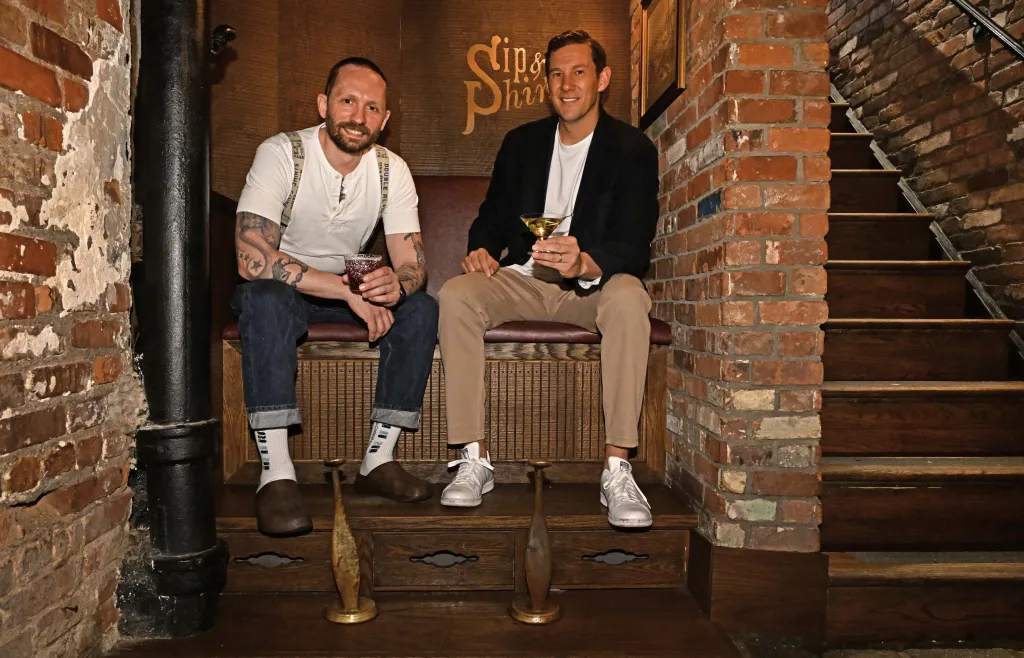 Two men sitting on a bench with cocktails, smiling at the camera.