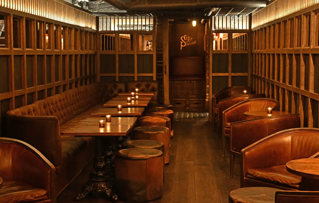 A dimly lit bar interior with booths, tables, and lounge seating, with a 