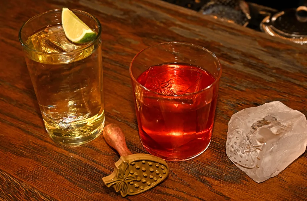 Two cocktails on a wooden bar with a bronze strainer and a large ice cube.