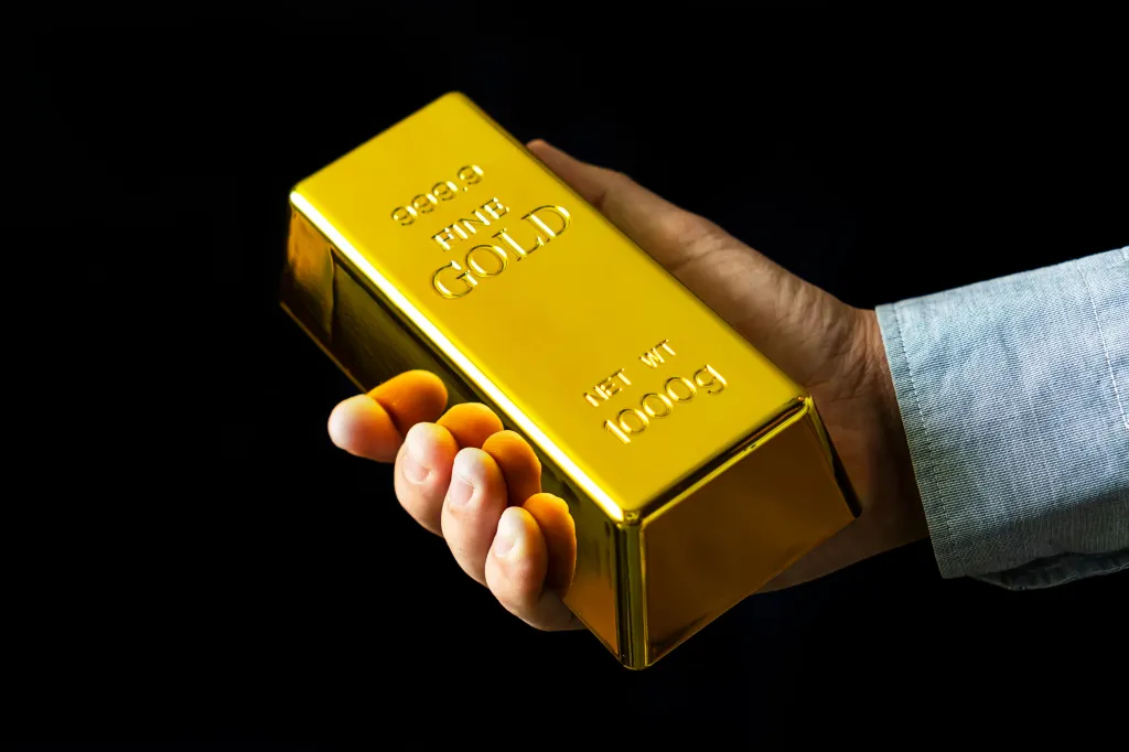 A hand in a blue shirt sleeve holds a 1000g fine gold bar against a black background.