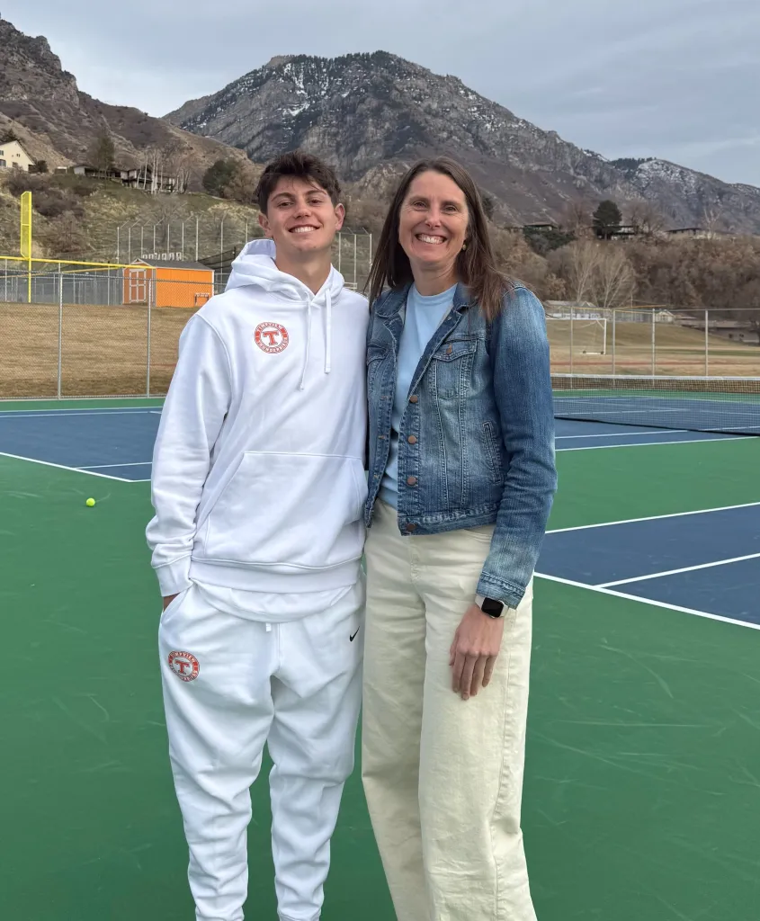 A young man and woman stand on a tennis court with mountains in the background.