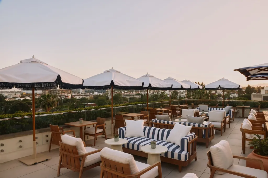 Rooftop patio with white umbrellas, striped couches, and wooden chairs overlooking a city.