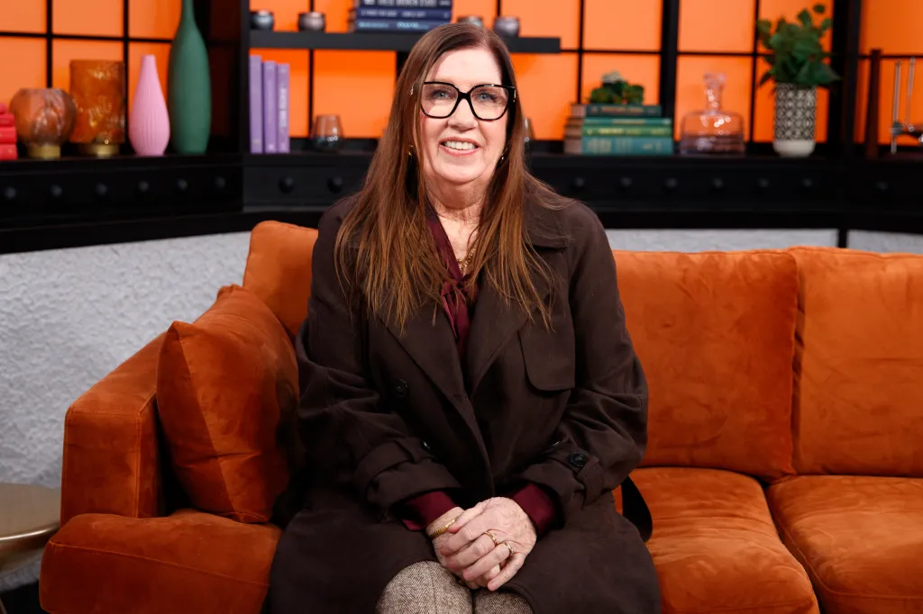 Jenny Slatten smiling while sitting on an orange couch, wearing a dark brown trench coat.