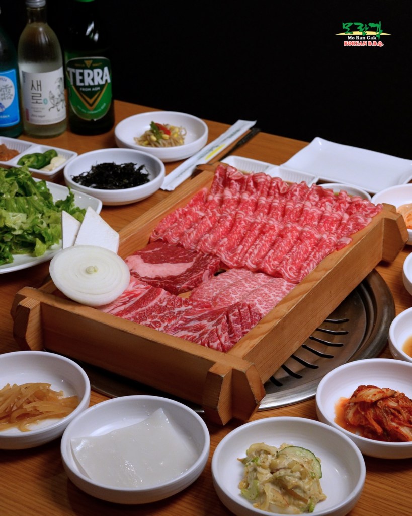 A variety of raw meats, side dishes, and drinks on a table at Mo Ran Gak Korean BBQ.