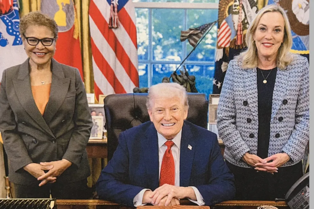 President Trump smiles while sitting at his desk, flanked by Congresswoman Karen Bass and another woman.