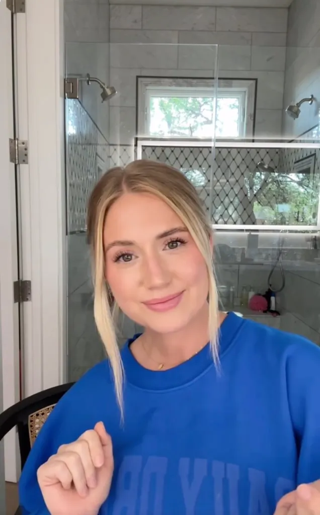 A woman in a blue sweatshirt smiles at the camera in a bathroom.