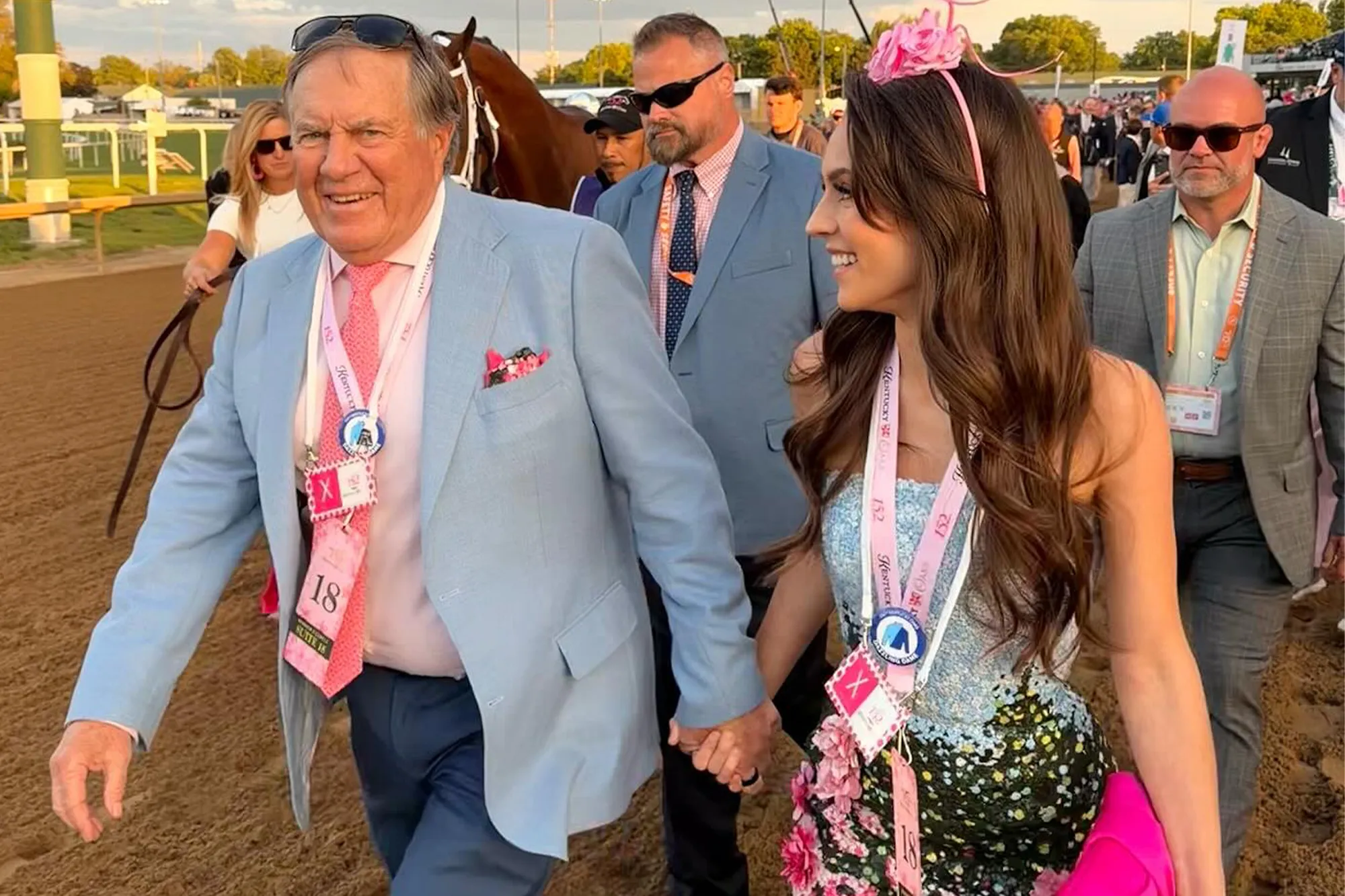 Bill Belichick and Jordon Hudson in formal attire hold hands while walking on a dirt track, with other people and a horse in the background.