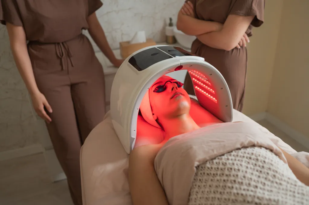Female patient undergoing red light phototherapy on her face and neck, with two female estheticians in the background.