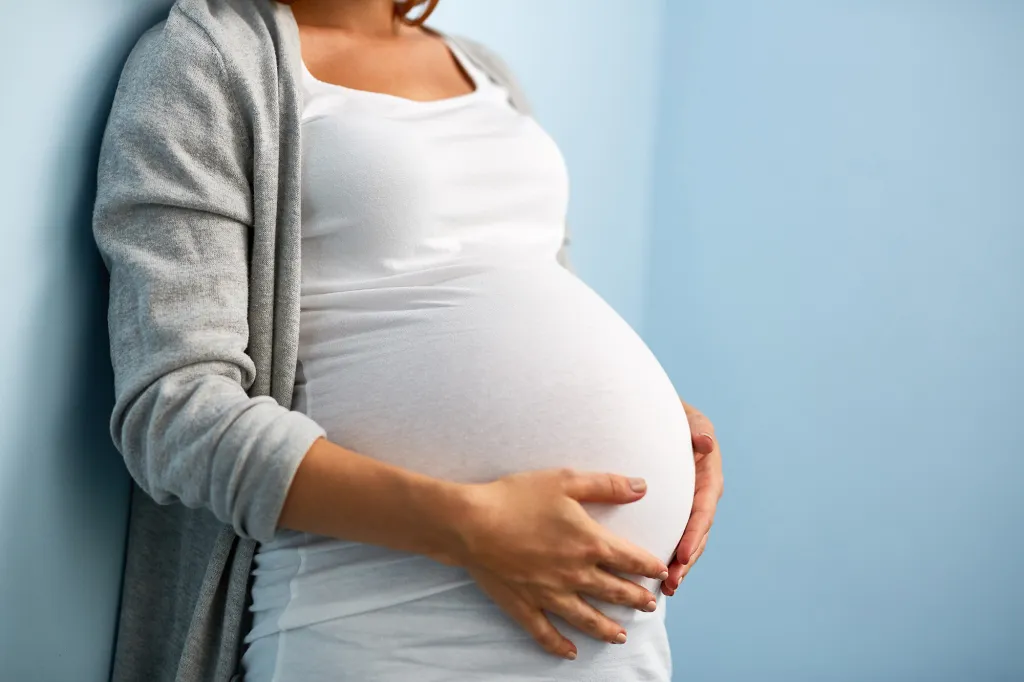 Pregnant woman in a white shirt and gray cardigan holding her belly.