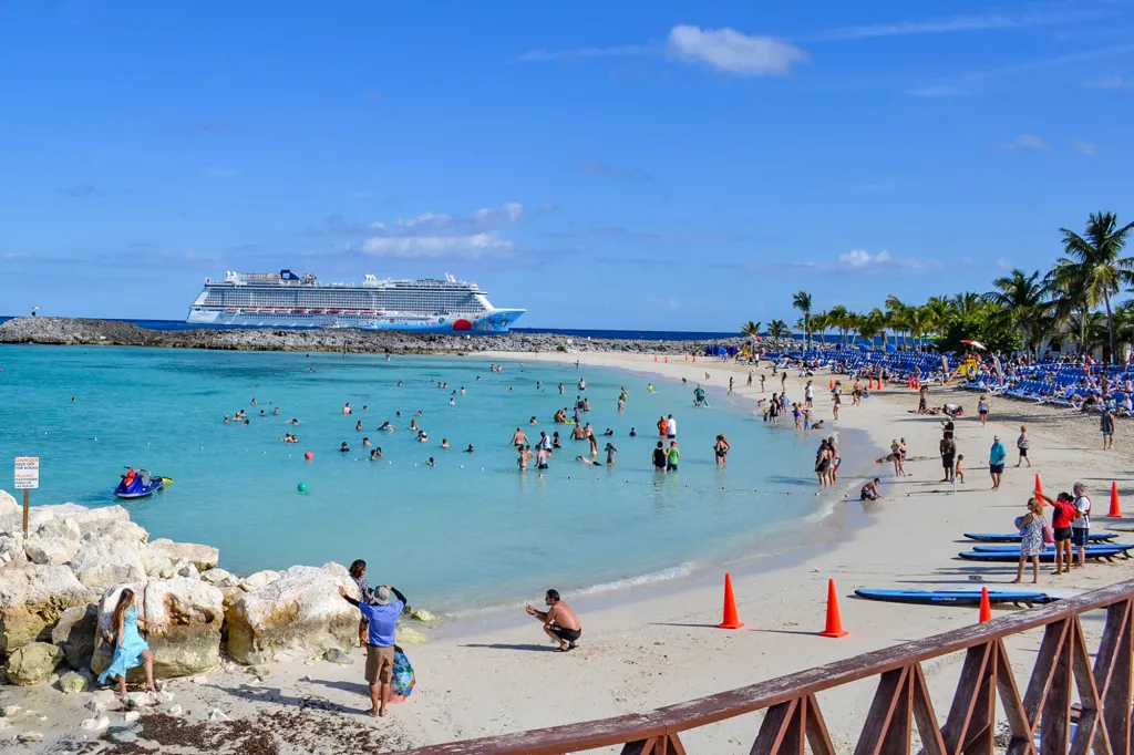 A cruise ship docked near a crowded beach with many people swimming and relaxing.