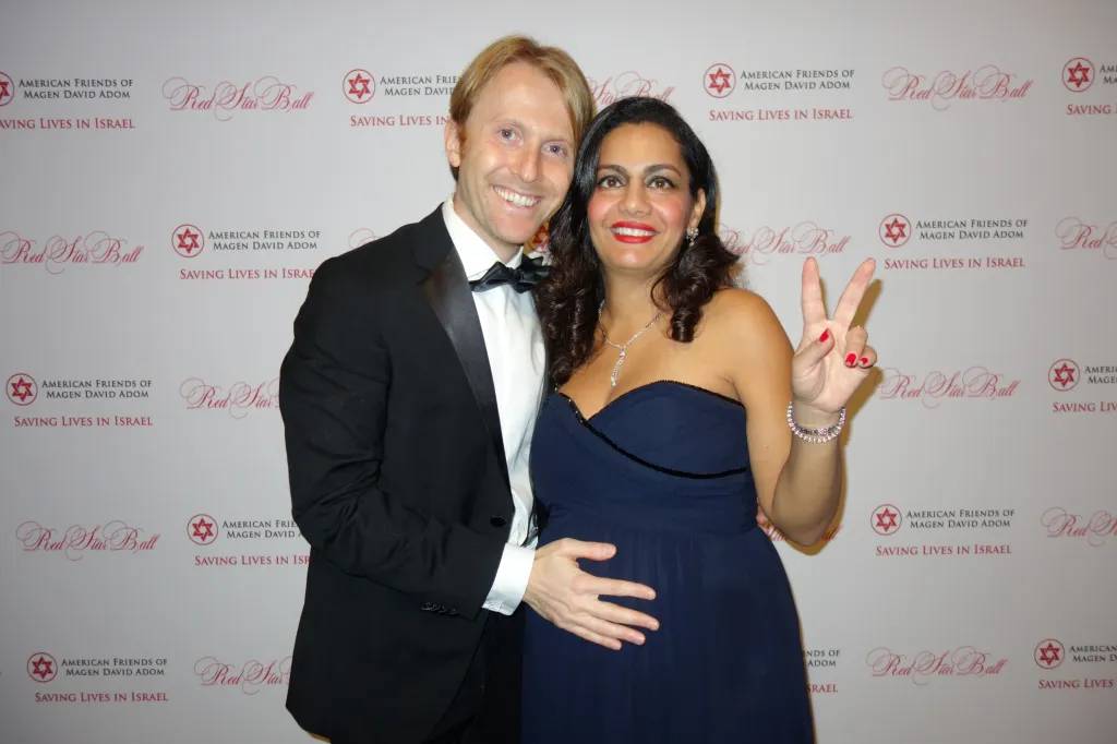 A man in a tuxedo with his arm around a pregnant woman in a navy dress who is making a peace sign.