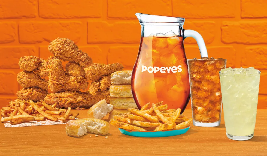 A Popeyes feast of fried chicken, fries, biscuits, a pitcher of iced tea, and drinks.