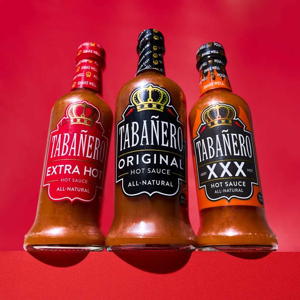 Three bottles of Tabañero hot sauce, including Extra Hot, Original, and Very XXX Hot varieties, on a red background.
