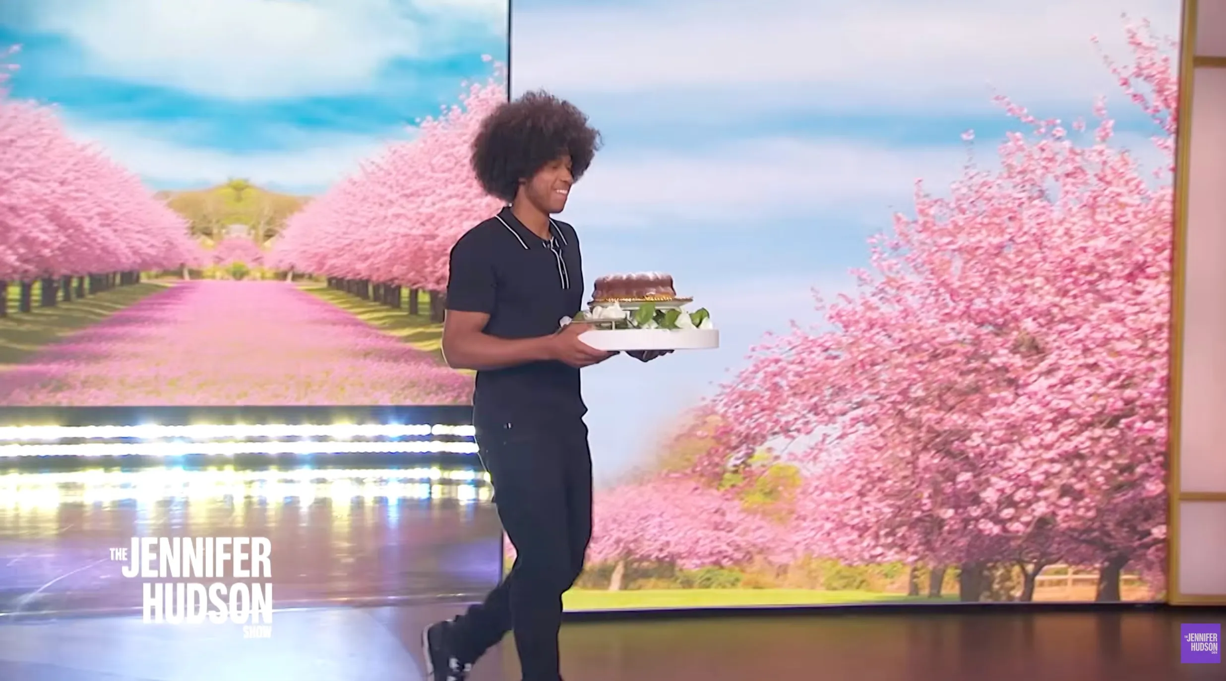 A man carries a cake on a tray while walking on a stage with a backdrop of pink cherry blossom trees.
