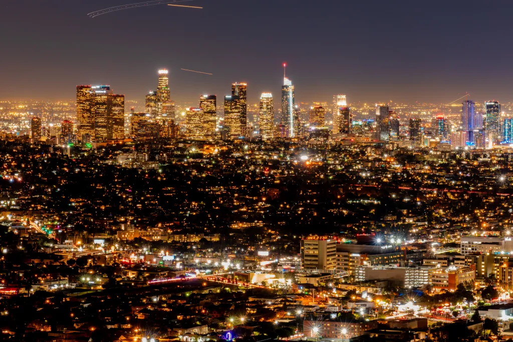 Wide night view of the glowing Downtown Los Angeles skyline with illuminated skyscrapers and dense city lights across the metropolitan area.