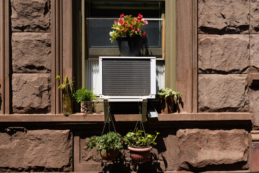A New York City apartment window with an air conditioner and potted plants.