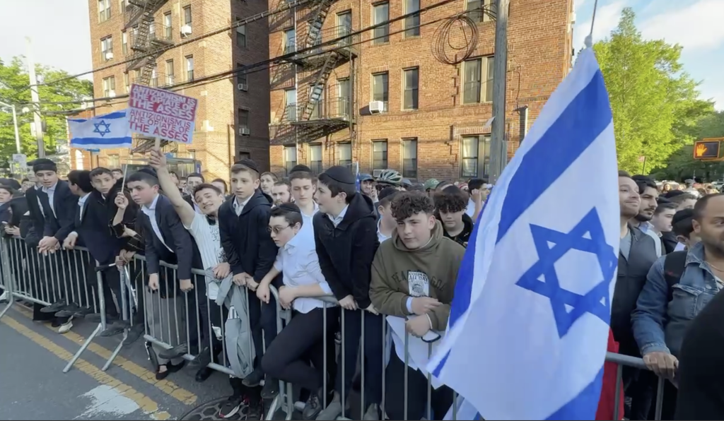 Protesters supporting Israel and Palestine near the Young Israel Synagogue of Midwood, with one person holding an Israeli flag next to a bus.