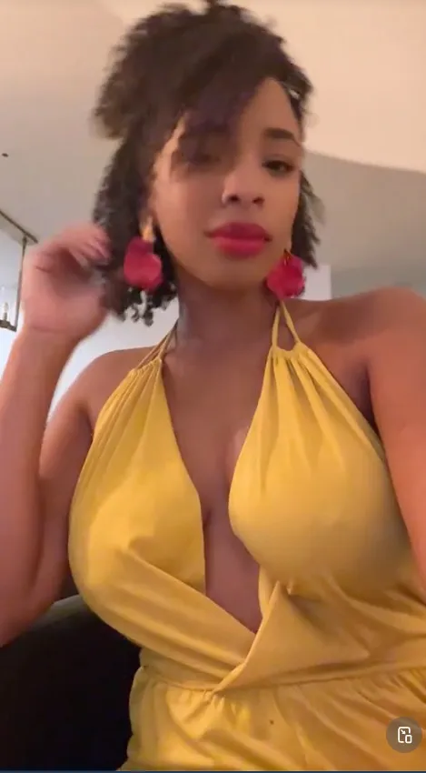 A woman with dark curly hair, wearing a yellow low-cut top and pink earrings.