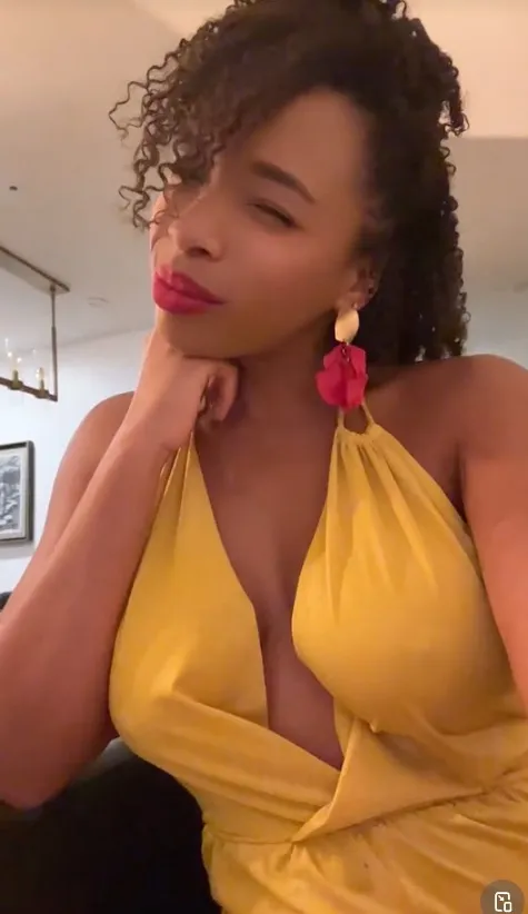 A woman with curly brown hair, wearing a yellow dress and pink earrings, looking at the camera with her chin resting on her hand.