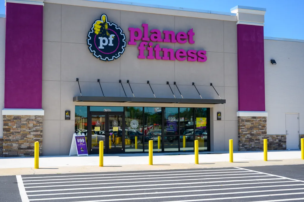 Exterior view of a Planet Fitness gym with purple and beige walls, glass doors, and a sign advertising a 
